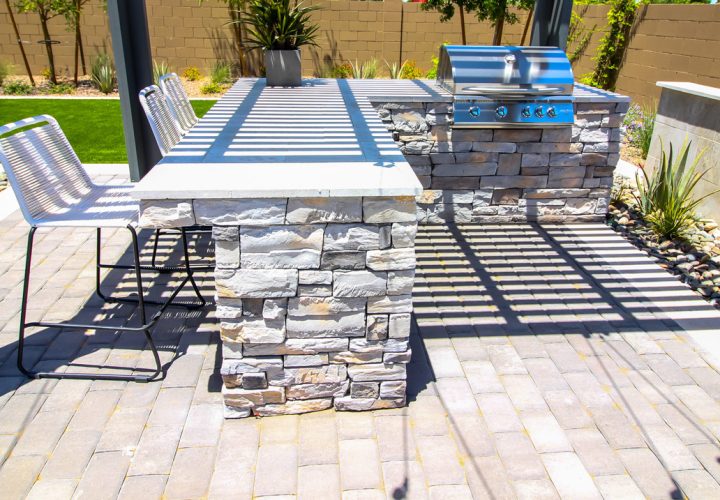 A modern outdoor kitchen setup with a stainless steel grill built into a stone-clad island, accompanied by a matching stone bar counter and metal bar stools. The area is set on a paver patio with landscaping and a covered pergola overhead, perfect for entertaining.