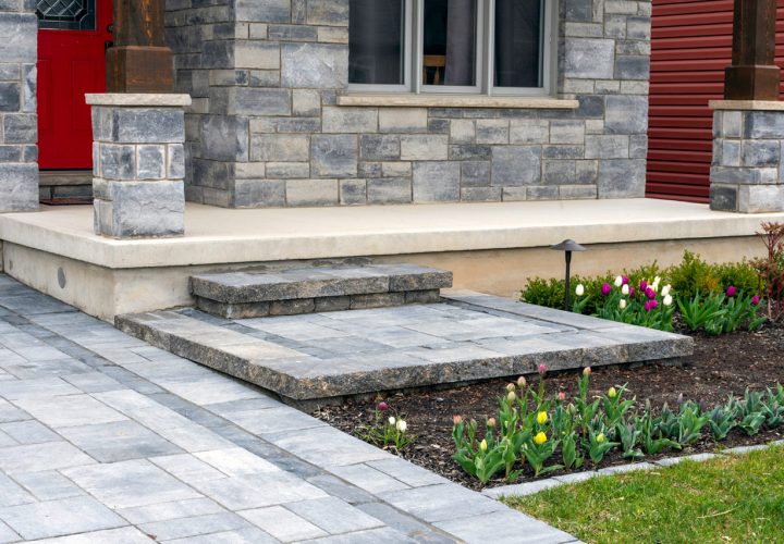 A clean, modern front porch featuring a smooth concrete landing with stone steps leading up to a red door. The entry is framed by gray stone pillars and flanked by landscaped flower beds with colorful tulips, all connected by a paved walkway for a welcoming and elegant approach.