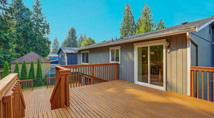 A modern elevated wooden deck with rich-toned planks and matching railings, attached to a gray-sided house with a glass sliding door. The deck overlooks a serene neighborhood with evergreen trees, providing a peaceful outdoor retreat.