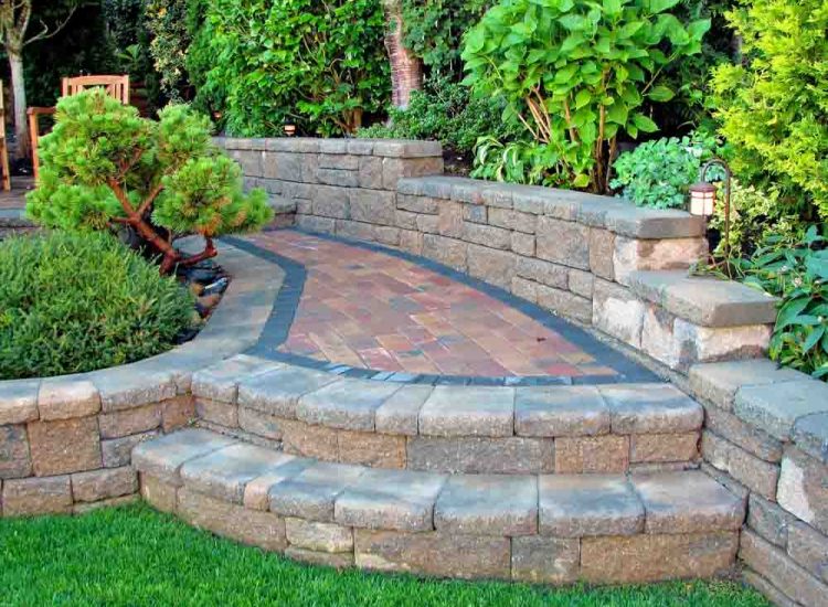 A beautifully designed curved retaining wall made of natural stone, enclosing a paver pathway with a decorative border. The area is landscaped with green shrubs, small trees, and a lantern-style light fixture, creating a tranquil garden setting.