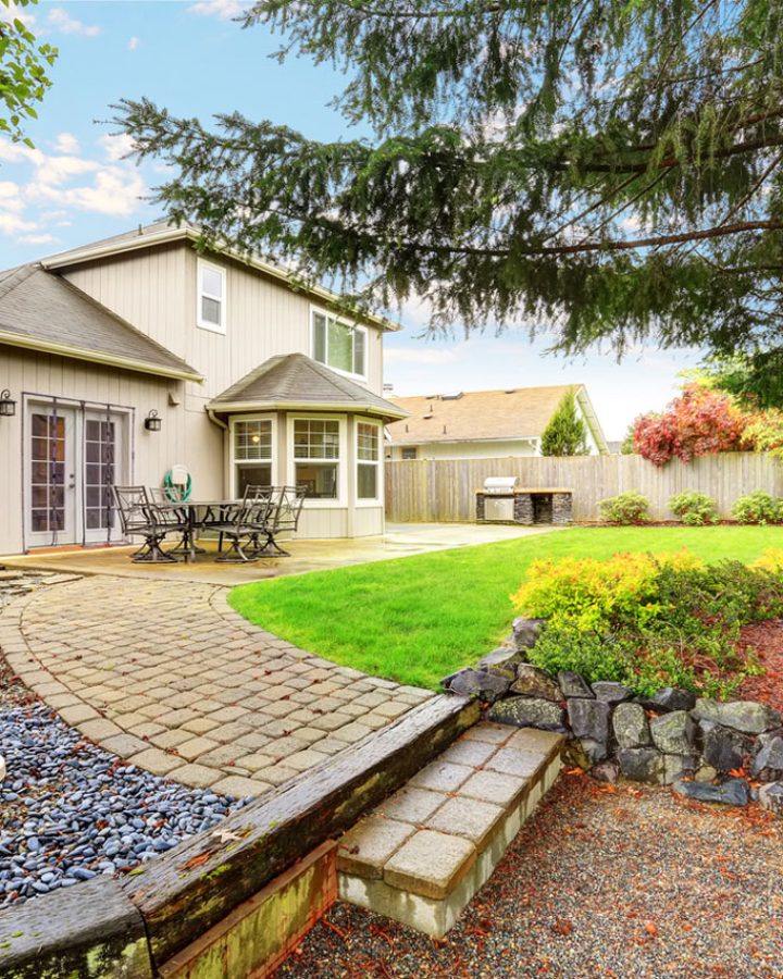 A well-designed backyard with a winding paver path leading through a mix of gravel, mulch, and rock gardens. The space includes a birdbath, flower beds, a patio area, and mature trees, offering a harmonious blend of hardscaping and natural beauty.