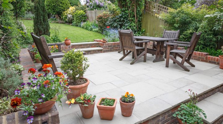 A charming stone patio furnished with wooden chairs and a table, bordered by colorful potted flowers and greenery. The space features a raised brick edge, steps leading to a larger lawn, and a backdrop of blooming shrubs and trees, creating a peaceful and inviting garden oasis.