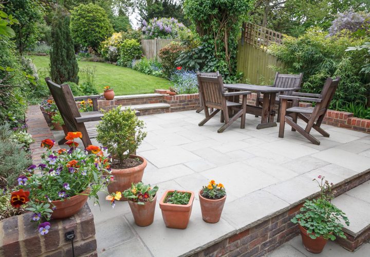A charming stone patio furnished with wooden chairs and a table, bordered by colorful potted flowers and greenery. The space features a raised brick edge, steps leading to a larger lawn, and a backdrop of blooming shrubs and trees, creating a peaceful and inviting garden oasis.