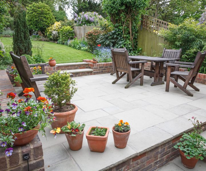 A charming stone patio furnished with wooden chairs and a table, bordered by colorful potted flowers and greenery. The space features a raised brick edge, steps leading to a larger lawn, and a backdrop of blooming shrubs and trees, creating a peaceful and inviting garden oasis.