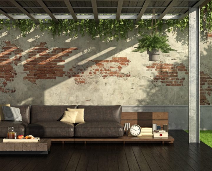 A cozy outdoor lounge area under a wooden pergola with hanging greenery, featuring a leather sofa, wooden coffee table, and exposed brick wall backdrop. The space includes a bicycle, potted plants, and a fruit-bearing tree, creating an inviting and rustic ambiance for relaxation.